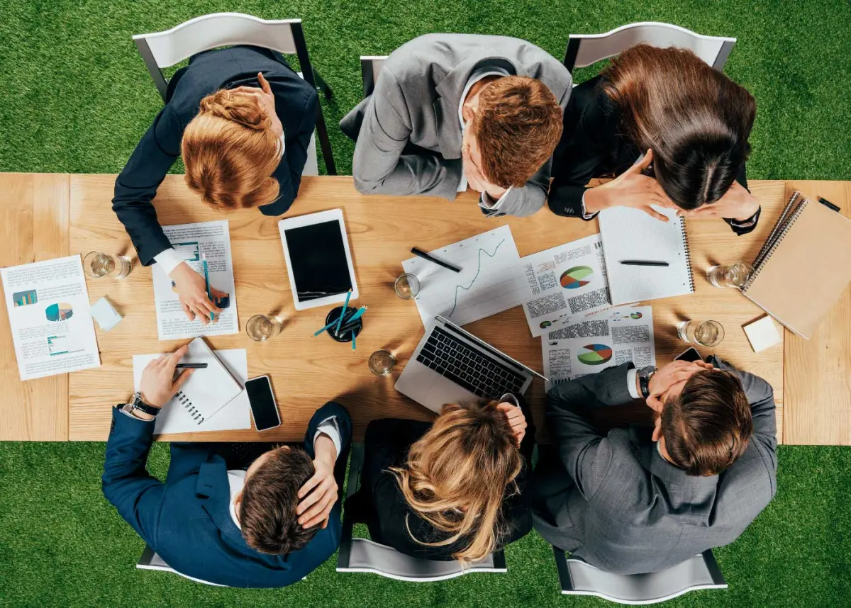 Blick von oben auf eine Beratungssituation: mehrere Personen sitzen an einem Tisch mit Unterlagen, Diagrammen, Laptop und Tablet und arbeiten zusammen.